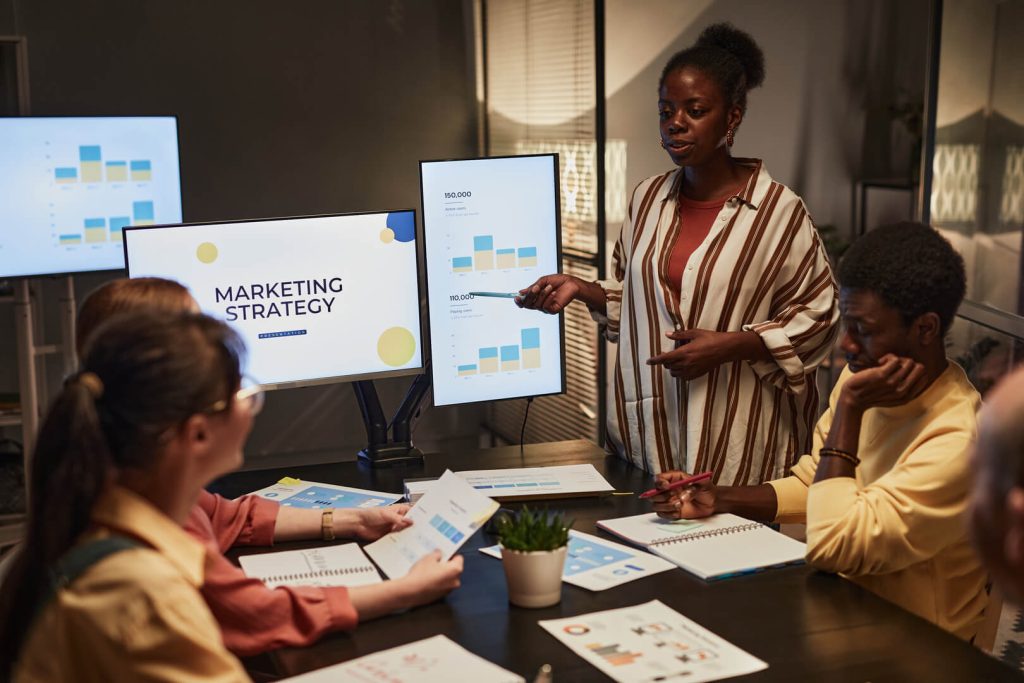 A woman is presenting a chart to a group of people seated around a table, illustrating the use of the 5W1H approach in strategy planning. Each participant appears engaged, some taking notes and others looking directly at the presenter. The room is well-lit, suggesting a professional environment.