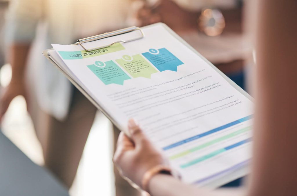 Close-up view of a paper filled with marketing analytics data, including graphs, charts, and numerical analyses. The paper is on a wooden desk surrounded by a pen, a cup of coffee, and some notes, indicating an ongoing analysis or work session.