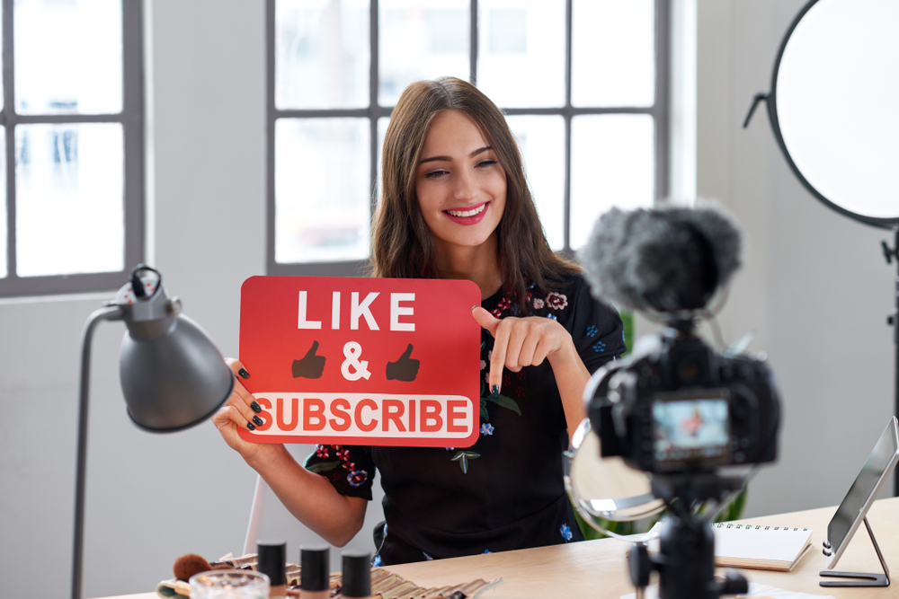 Female content creator recording a video while holding a 'Like & Subscribe' sign—illustrating a common and effective tactic in how to promote a podcast by encouraging audience engagement across platforms.