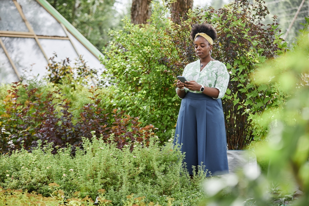 Woman in garden using smartphone to manage tree service leads, surrounded by lush greenery and trees.