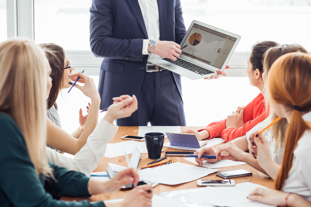 Team members discussing a local marketing strategy during a meeting, with one person presenting data on a laptop showing pie charts, while others seated around a table take notes and engage in conversation.