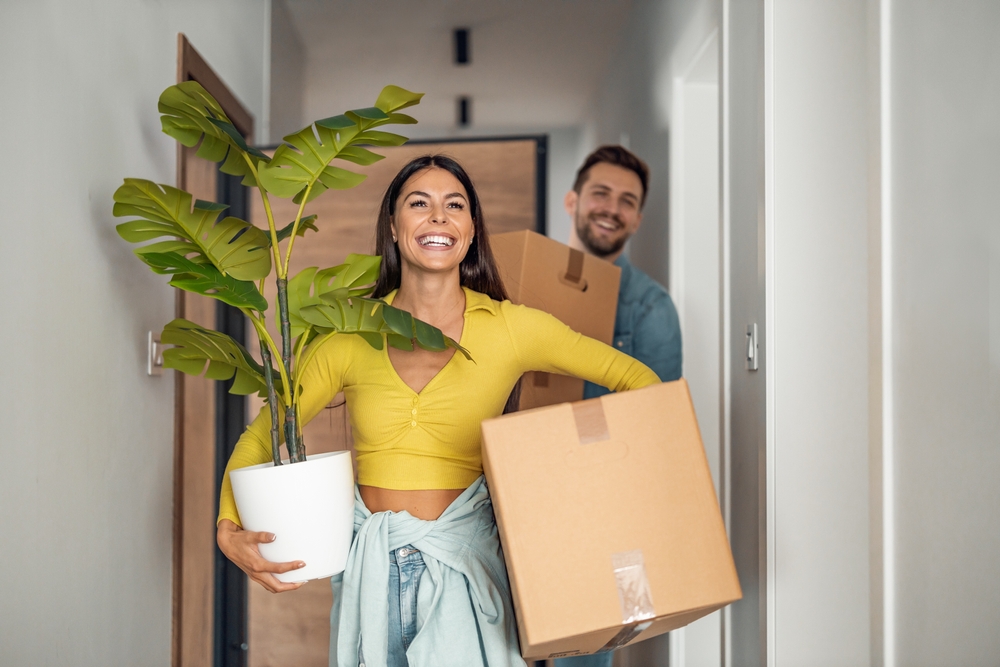 Happy couple carrying boxes and a houseplant into a new home, symbolizing a fresh start—ideal for moving company advertising focused on stress-free and joyful relocation experiences.