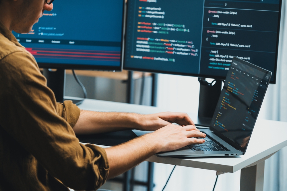 A developer working on code at a standing desk with multiple screens displaying programming scripts. The image highlights developer marketing themes by showcasing modern coding, technology use, and productivity.