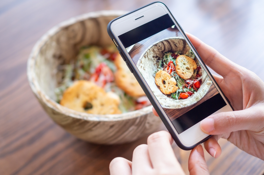 A person taking a photo of a delicious salad with croutons in a rustic bowl, emphasizing the importance of high-quality food images for restaurant marketing.
