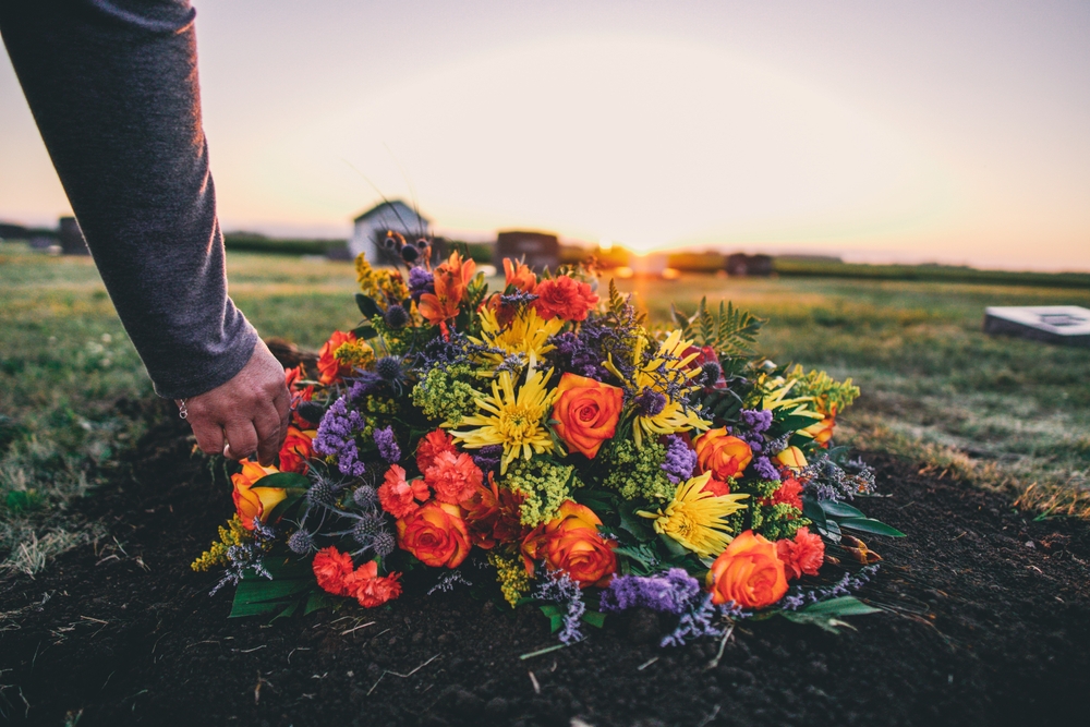 A hand places a colorful funeral flower arrangement on a fresh grave at sunset — symbolic image for how to market a funeral home with compassion and care.