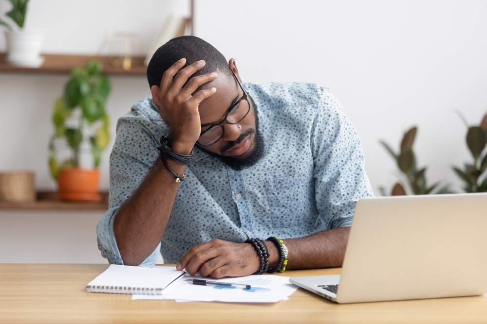 Frustrated man sitting at a desk with his hand on his head, staring at marketing charts and a laptop, humorously representing the stress and lessons learned from funny marketing fails.