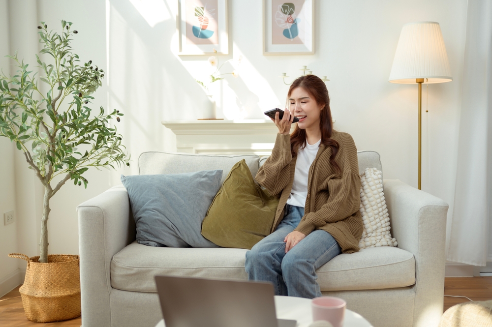 Woman sitting on a sofa using voice search on her smartphone, illustrating human search behavior patterns as users rely on hands-free, conversational queries.