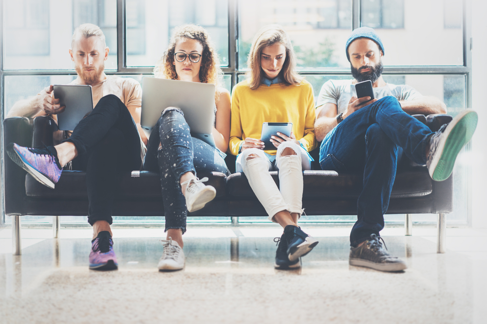 A group of young professionals sitting on a couch using laptops, tablets, and smartphone, symbolizing a modern outsourced digital marketing team collaborating remotely.