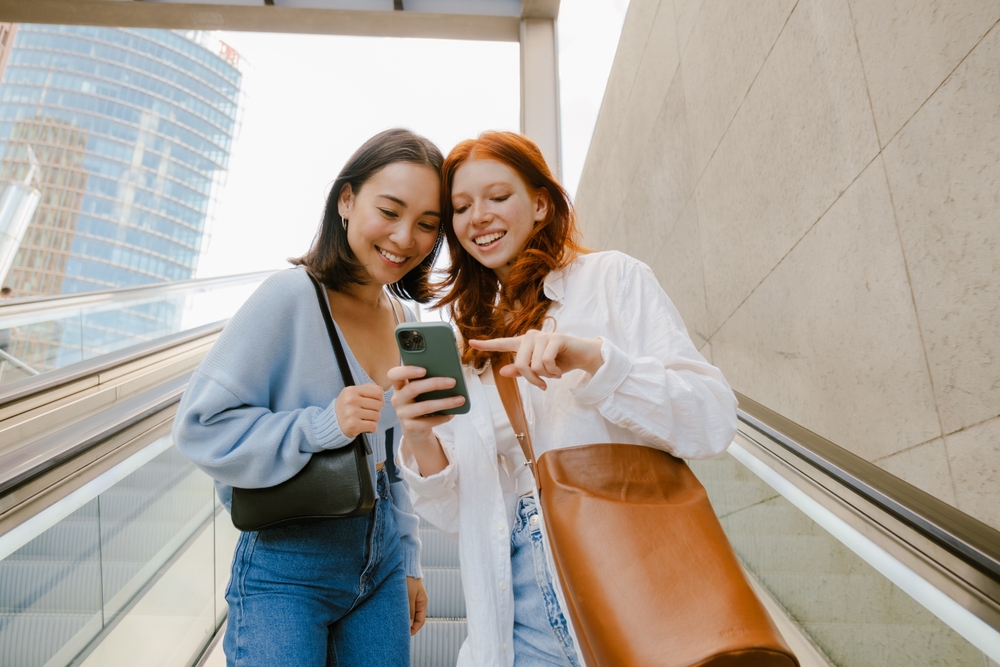 Two young women on an escalator smiling as they watch a clip on a smartphone, enjoying content produced by an advertising video company.