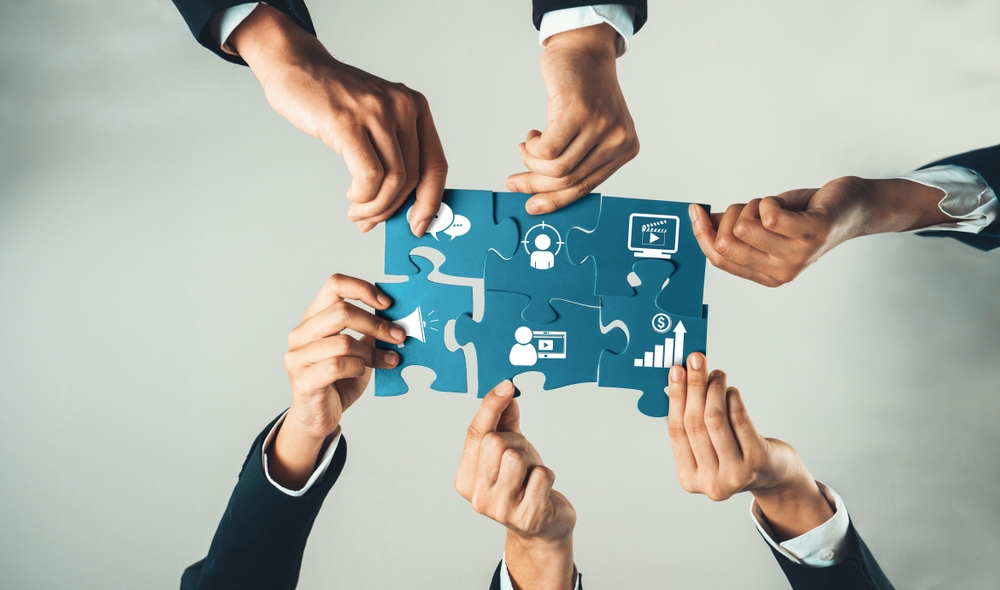 Multiple hands assembling puzzle pieces with marketing icons, symbolizing how teams collaborate to solve marketing bottlenecks and streamline strategy.