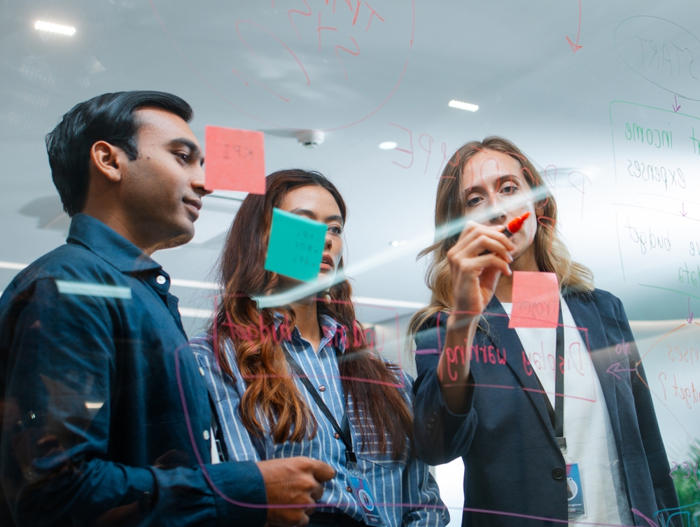 Three professionals collaborate at a glass whiteboard, illustrating teamwork, innovation, and thought leadership in a modern office.