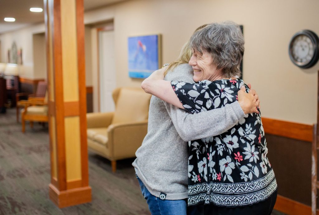 A resident and a caregiver share a hug in the lounge of a senior living community
