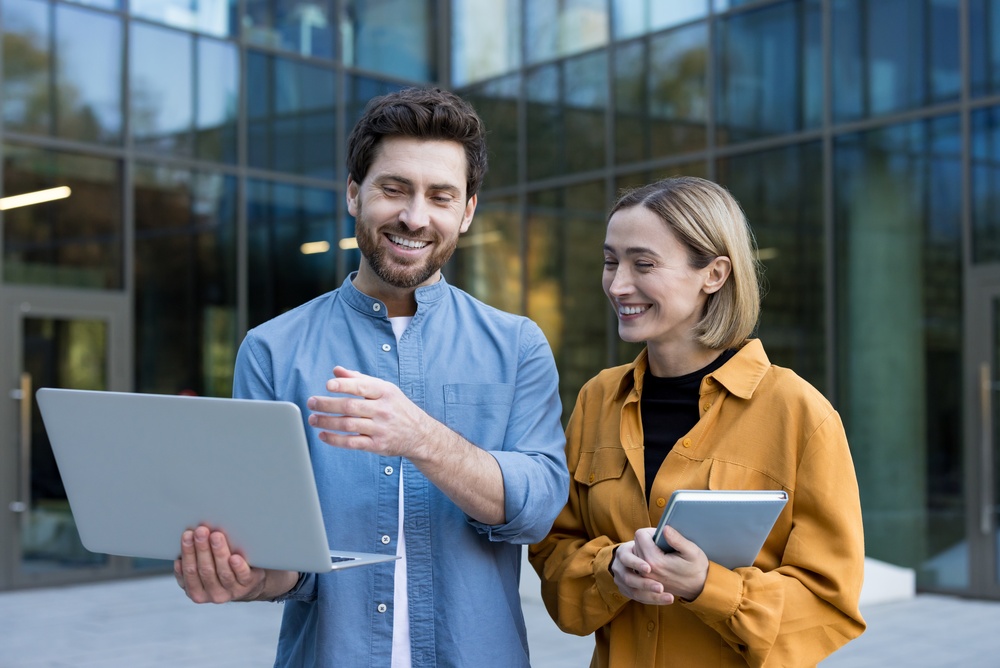 Two professionals reviewing a laptop outside a modern office building, representing collaborative B2B content strategy and workplace communication.