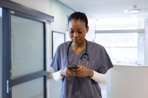 Nurse in gray scrubs with a stethoscope checking her phone in a hospital corridor