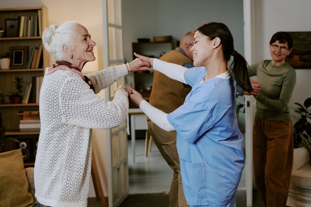 Young caregiver dancing with a smiling senior woman while two residents look on, an unscripted scene for assisted living marketing ideas.