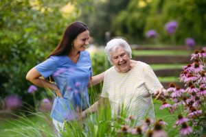 Caregiver in blue scrubs laughing with an elderly woman in a flowering garden, the kind of real moment assisted living marketing should show.