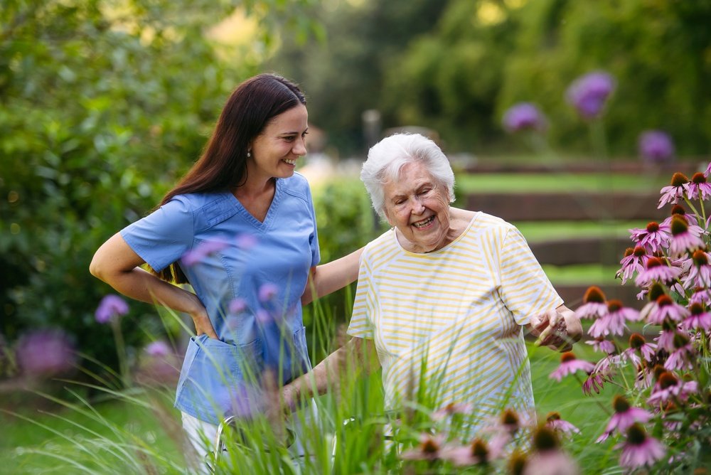 Caregiver in blue scrubs laughing with an elderly woman in a flowering garden, the kind of real moment assisted living marketing should show.