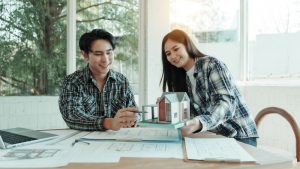 Smiling couple reviewing a house model and floor plans at a sunlit table while comparing home improvement contractors