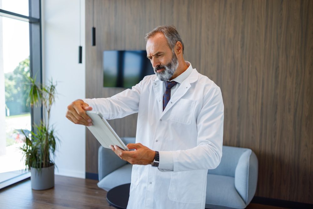 Doctor in a white coat holding a tablet at arm's length and reading it with a frown